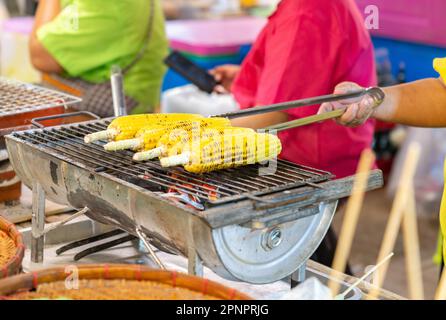 Woman grilled corn cobs and flip on grill in street food market. Stock Photo