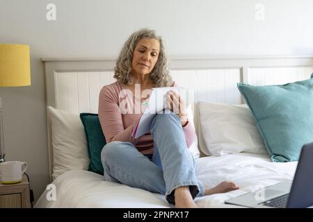 Happy senior caucasian woman sitting on bed and writing in notebook Stock Photo