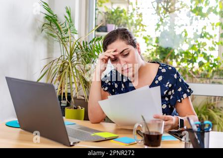 A young businesswoman sitting in her office desk, looking intently at paperwork and appearing to be feeling overwhelmed Stock Photo