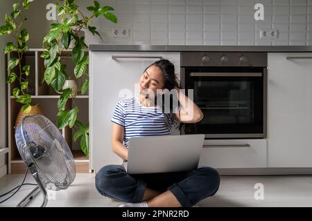 Contented Asian woman turn on floor ventilator fan while work with laptop joyful self save from heat Stock Photo