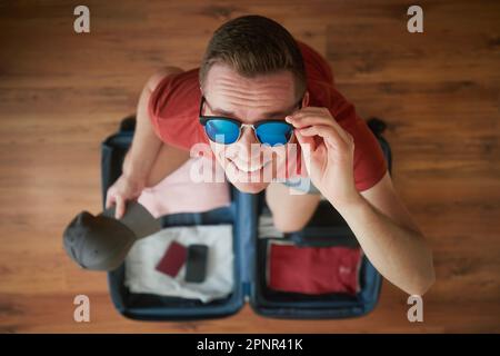 Packing suitcase and planning travel summer adventure. Portrait of happy smiling man with sunglasses looking at camera above open baggage. Stock Photo
