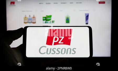 Person holding cellphone with logo of British consumer goods company PZ Cussons plc on screen in front of webpage. Focus on phone display. Stock Photo