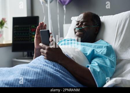 African American man smiling while talking online with his relatives on smartphone, he lying on bed in ward Stock Photo