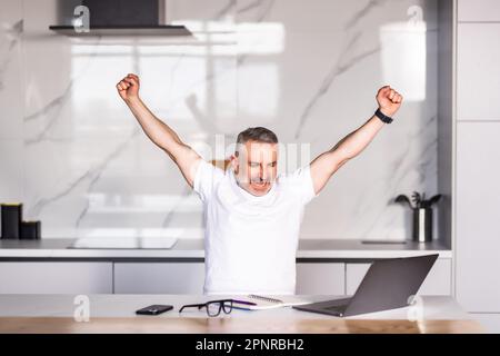 Excited man in glasses sit at table in kitchen feel euphoric win online lottery on laptop, overjoyed young male in spectacles look at computer screen Stock Photo