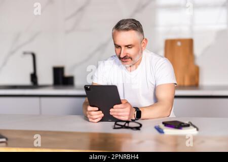 Middle-aged professional man looking at camera and checking messages on digital tablet while having breakfast on kitchen counter at home Stock Photo