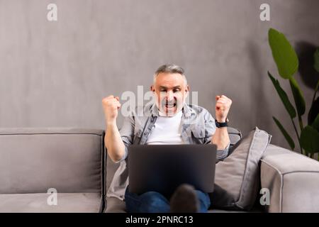 Emotional middle aged man in eyewear looking at computer screen, making thumbs up gesture feeling excited reading email with good news, celebrating on Stock Photo