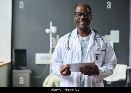 Portrait of African American male doctor in white coat smiling at camera while using digital tablet in ward Stock Photo