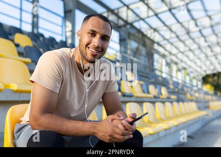 Portrait of a young African-American male sportsman sitting in the stands of the stadium, resting, listening to music in headphones from the phone. He looks at the camera, smiles. Stock Photo