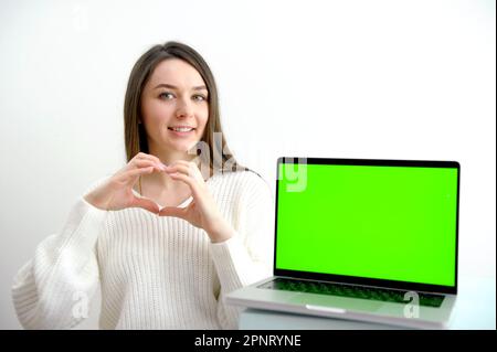Young woman speak with friends on video call and shows heart with hands on camera from home office. Virtual party meeting team teleworking. Remote conference Computer green screen view. Stock Photo