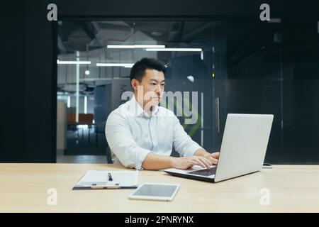 Portrait of a successful young Asian male programmer sitting in a modern office at a desk and working on a laptop. Stock Photo