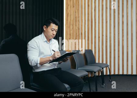 A young Asian man sits in the lobby and fills out a survey questionnaire in the medical center, in the office for work, interview, arrival at the hotel. Stock Photo