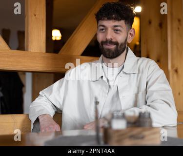 Young curly-haired man with a smile on the street sitting at a table in a restaurant in a wooden design Stock Photo