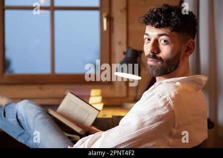 Young curly-haired man sitting at home with a book in his hands against the background of a window Stock Photo