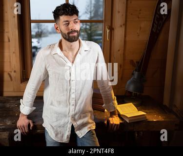 A young curly-haired man stands in the room against the background of the window, leaning on a wooden table Stock Photo
