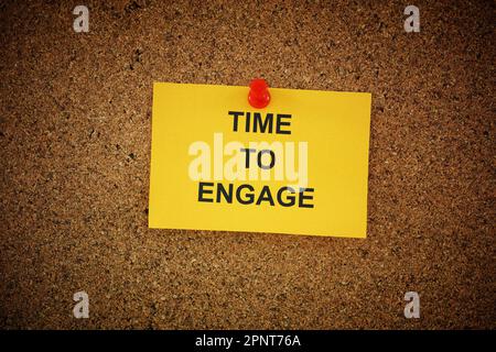 A yellow paper note with the words Time To Engage on it pinned to cork board. Close up. Stock Photo