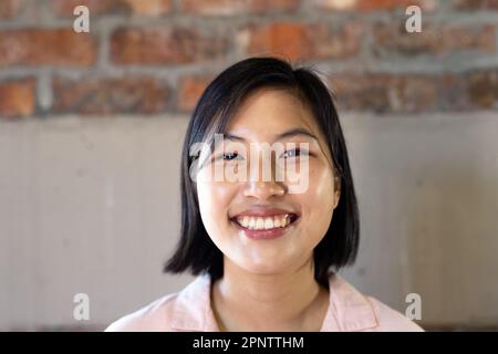 Portrait of happy asian casual businesswoman smiling against brick wall in office Stock Photo