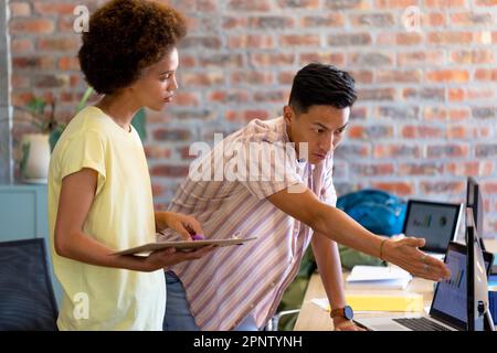 Two diverse creative colleagues using tablet and computer discussing project in casual office Stock Photo