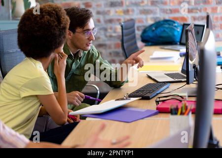 Two diverse creative colleagues using computer discussing project at desk in casual office Stock Photo