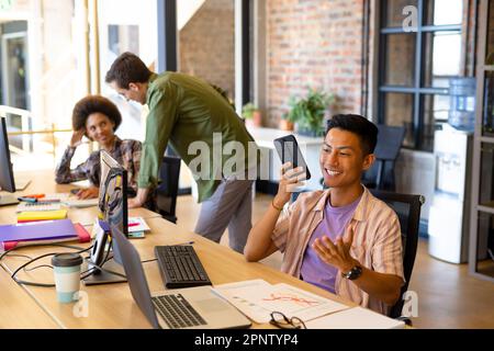 Happy asian male designer talking on smartphone at desk in creative office with diverse colleagues Stock Photo