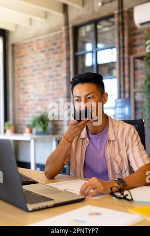 Happy asian male designer talking on smartphone and using laptop at desk in creative office Stock Photo