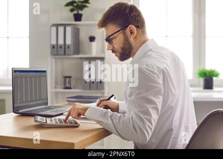 Young focused serious business man working on laptop at office making calculations. Stock Photo