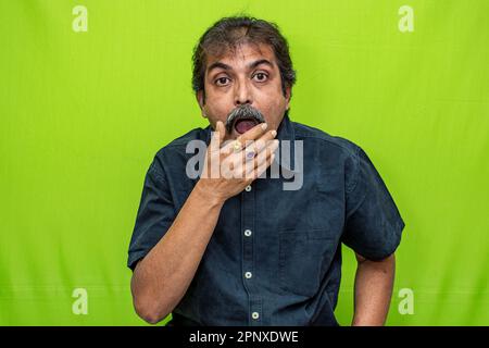 A well-dressed corporate man in a black shirt is seen standing against a green screen, with his right hand on his mouth in a surprised expression Stock Photo