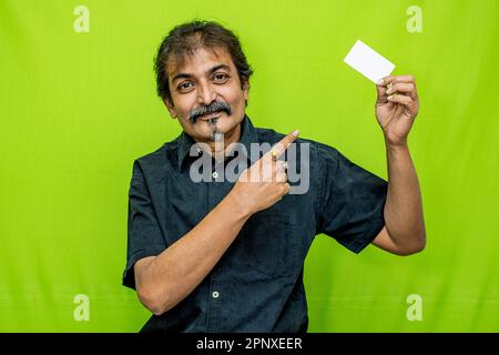 Corporate man in black shirt, smiling with a blank card in left hand and pointing with right, in front of green screen background Stock Photo