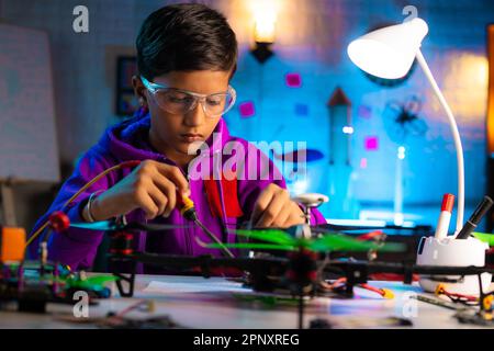 Intelligent serious indian kid busy making drone connections by soldering at home during night - concept of genius, smart kid and innovation. Stock Photo