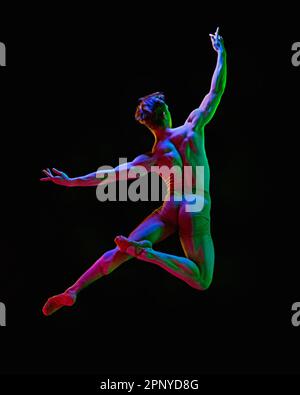 Portrait of graceful muscled male ballet dancer jumping up with spreaded hands over dark studio background with neon light. Levitate, back view Stock Photo