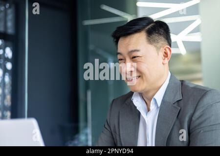 Closeup of cheerful and successful businessman smiling and looking at laptop screen, successful asian boss entrepreneur inside office at workplace looking at laptop screen, reading good news Stock Photo