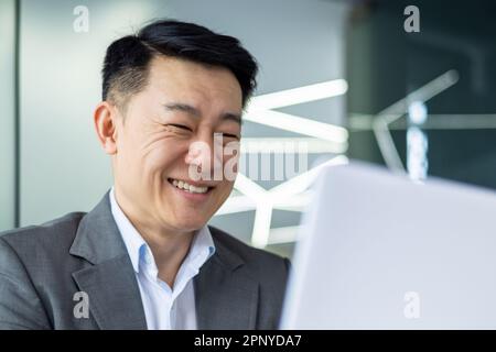 Closeup of cheerful and successful businessman smiling and looking at laptop screen, successful asian boss entrepreneur inside office at workplace looking at laptop screen, reading good news Stock Photo