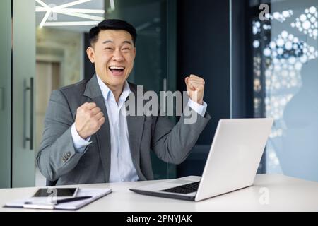 Portrait of successful asian boss, businessman celebrating results achievement, man in business suit smiling and looking at camera holding hands up triumph gesture, mature investor inside office. Stock Photo