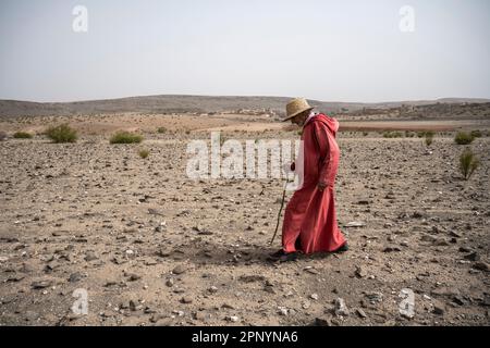Portrait of an elderly shepherd dressed in a red djellaba herding his flock in the arid lands of Morocco. Stock Photo