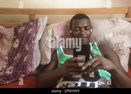 Handsome young African American man with wireless headphones listening to music and smiling while lying on bed and using his mobile phone Stock Photo