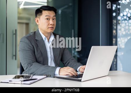 Surprised asian man looking at camera, mature adult boss businessman working inside office with laptop while sitting at workplace. Stock Photo