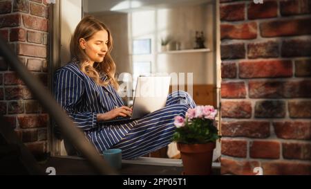 Caucasian Woman Sitting on her Windowsill, Using Laptop and Smiling. Female E-commerce Business Owner Happy About her Company's Growth as She Checks Stock Photo