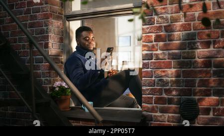 Black Authentic Man Using Smartphone in his Flat While Sitting on Bedroom Windowsill. African American Male Posting Photos on Social Media and Using Stock Photo