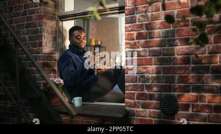 Black Authentic Man Using Smartphone in his Flat While Sitting on Bedroom Windowsill. African American Male Posting Photos on Social Media and Using Stock Photo