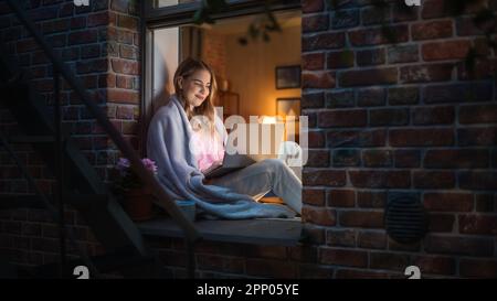 White Young Woman Working at Night with Laptop From the Comfort of her Home, Sitting on Windowsill. Female Client Advisor Answering Customer Support Stock Photo