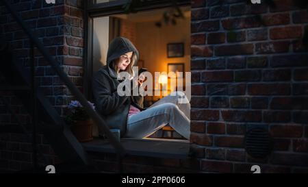 White Woman Wearing a Hoodie Using her Smartphone While Sitting on Windowsill at Night. Female Gamer Playing Addictive Mobile Phone Games Instead Stock Photo