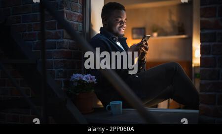 Black Man Using Smartphone for Online Shopping While Sitting on his Windowsill at Night. e-Commerce Concept of Easy Purchasing, Buying, and Ordering Stock Photo