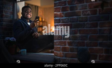 Black Man Using Smartphone for Online Shopping While Sitting on his Windowsill at Night. e-Commerce Concept of Easy Purchasing, Buying, and Ordering Stock Photo