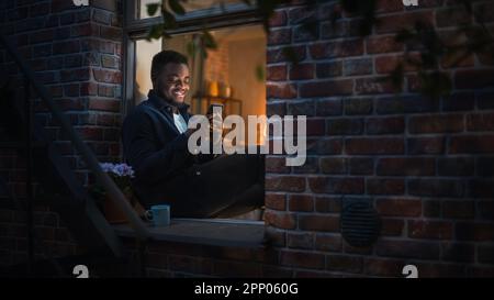 Black Man Using Smartphone for Online Shopping While Sitting on his Windowsill at Night. e-Commerce Concept of Easy Purchasing, Buying, and Ordering Stock Photo