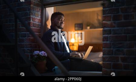 Young Handsome Black Man Working from Home on Laptop Computer in Windowsill of a Cozy Stylish Loft Apartment. Male Smiling, Checking Social Media Stock Photo