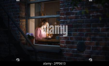Beautiful Blond Woman Works on a Laptop while Sitting at Her Desk in the Bedroom. Female Professional Freelancer Working from Cozy Home at Night. Stock Photo