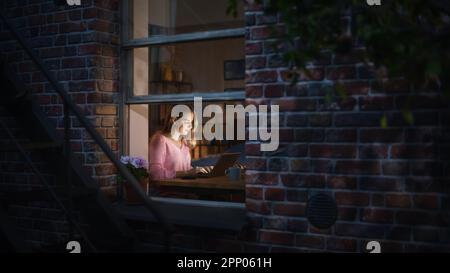 Beautiful Blond Woman Works on a Laptop while Sitting at Her Desk in the Bedroom. Female Professional Freelancer Working from Cozy Home at Night. Stock Photo