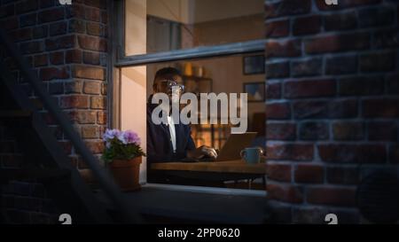 Portrait of Black Creative Young Man Working from Home on a Laptop in Cosy Warm Apartment During Night. African American Male Developer Engineering Stock Photo