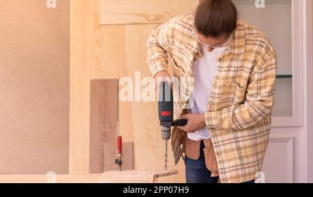 Concentrated young workman in checkered shirt using electric screwdriver while working with wood Stock Photo