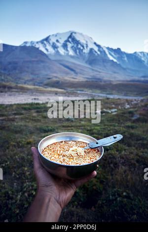Tourist holding bowl full of noodles fast food and spoon in hand in nature mountain valley camping in trekking of Central Asia, Kazakhstan Stock Photo