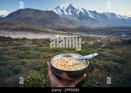 Tourist holding bowl full of noodles fast food and spoon in hand in nature mountain valley camping in trekking of Central Asia, Kazakhstan Stock Photo